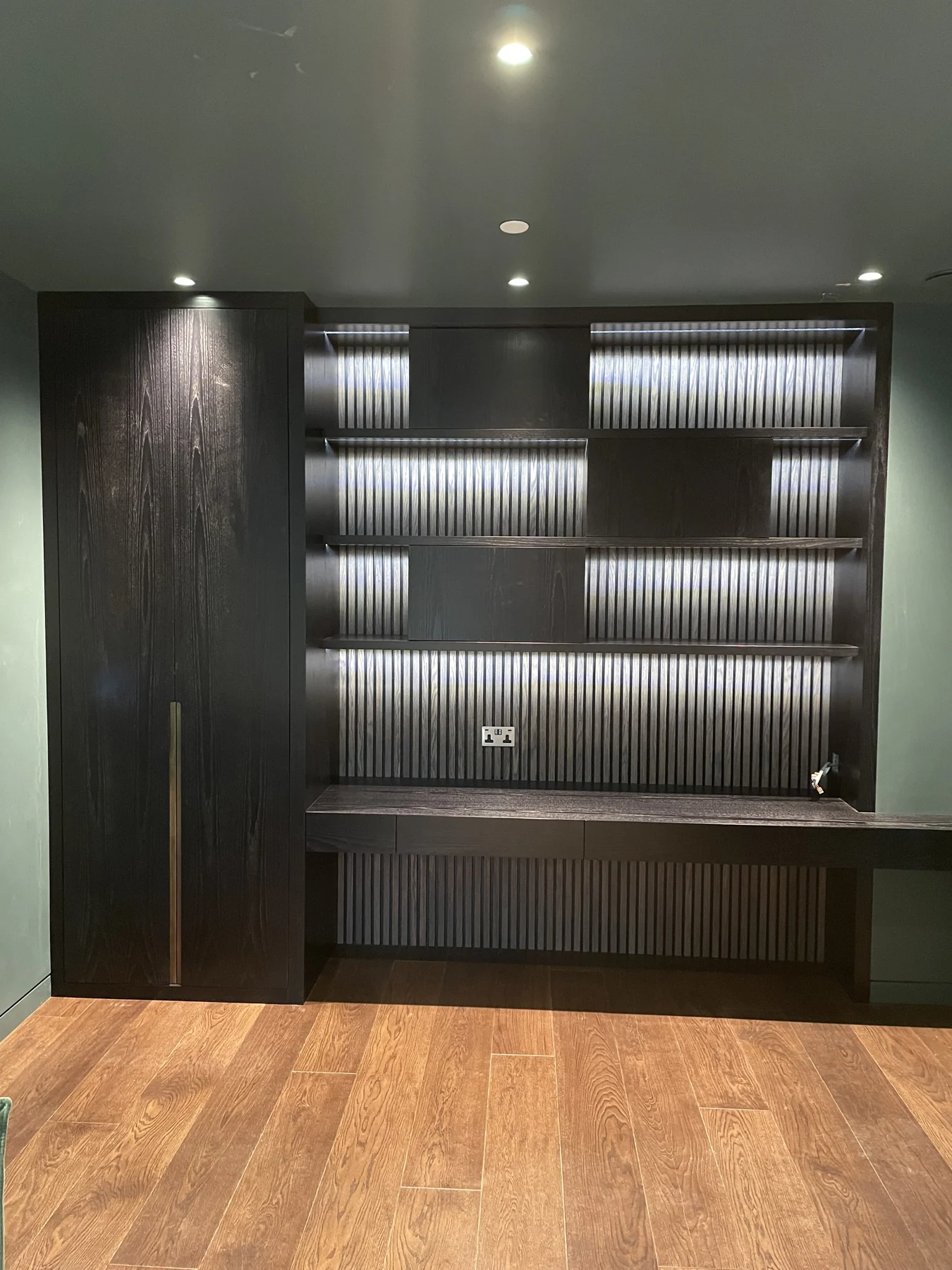 Dark shelving wall with ribbed backing, integrated lighting and a low bench-like cabinet.